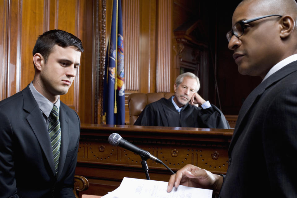 A man in a suit testifies in court, standing at a microphone. Another man in a suit holds documents while a judge listens attentively in the background