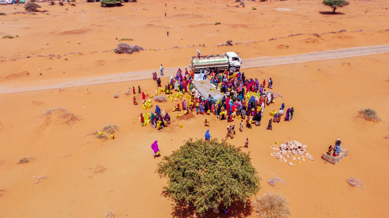 An aerial shot of a truck by a narrow road dispensing water from a giant bladder to dozens of women in brightly colored shawls.