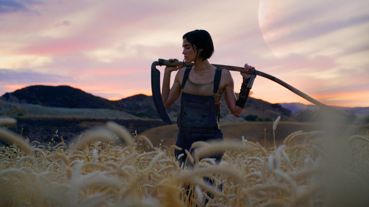  Kora stands in a field of wheat with a scythe over her shoulders in Rebel Moon Part 1. 