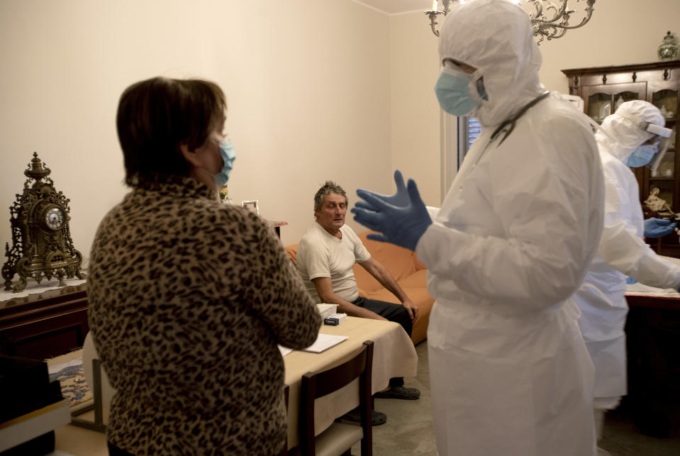 Doctor Luigi Cavanna, right, talks with COVID-19 patient Giancarlo Salvi's wife Luciana as he listens from the couch, in their home, in San Nicolo, near Piacenza, Italy, Wednesday, Dec. 2, 2020. (AP Photo/Antonio Calanni)