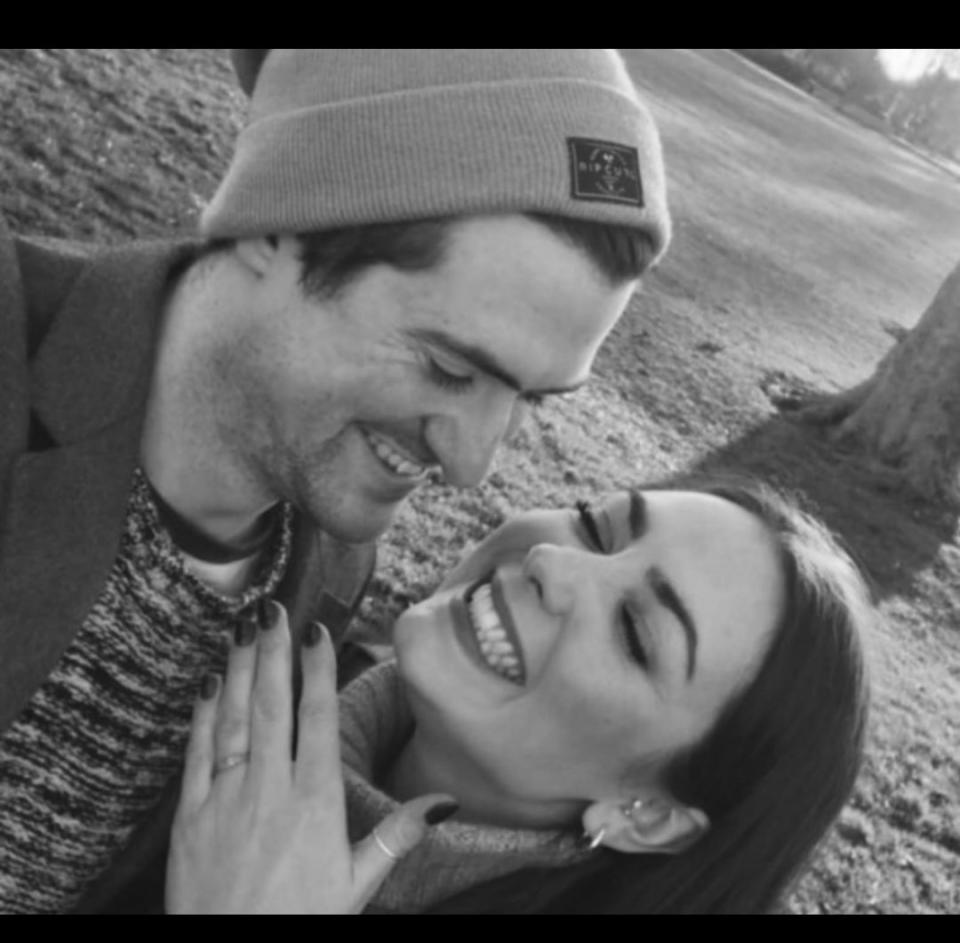 A man and woman pose for a selfie. The man (left) is wearing a beanie hat and looking at his fiance (right) who is holding her hand up to the camera