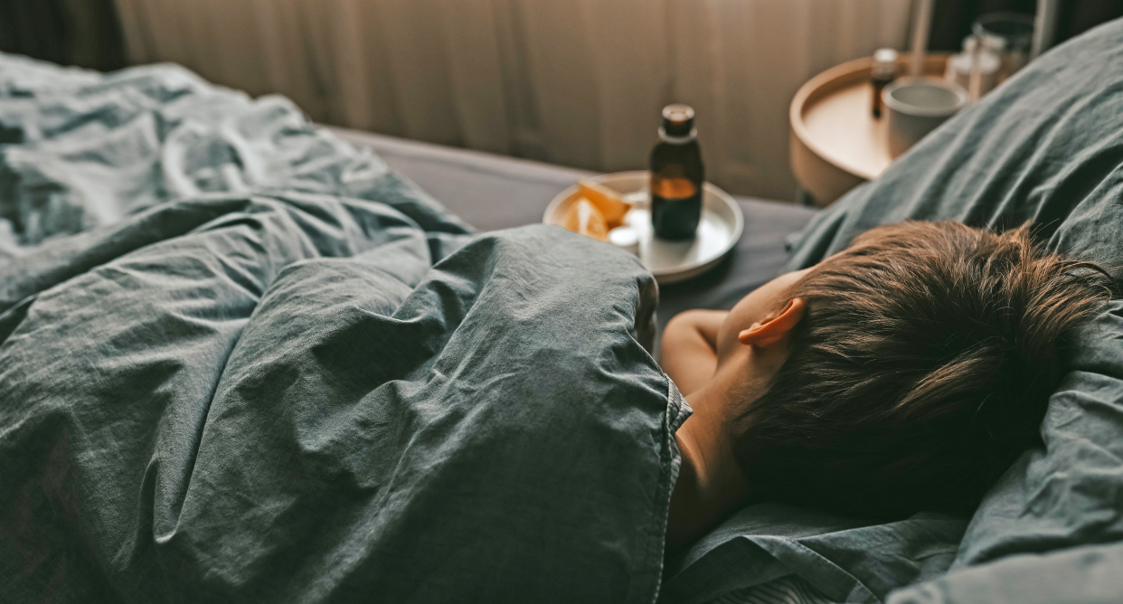 A young child with strep A sick in bed under dark gray covers, with medicine and two orange slices on a plate beside them.