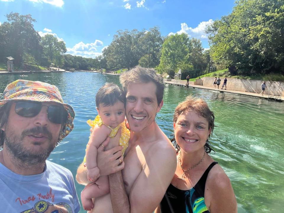 Photo of Ash Jurberg and his cousin Yonatan. Ash is on the left side of the photo and has salt and pepper hair, with a mustached and closed-cropped beard. He wears a colorful bucket hat, dark sunglasses, and a blue t-shirt. Yonatan has dark brown hair, blue eyes, and has no shirt on. He holds a baby to his cheek. The baby has dark brown hair, dark eyes, and wears a yellow dress with white polka dots. To the right of Yonatan is an older woman who has blue eyes and red-brown short hair that is wet. She wears a black one piece bathing suit and silver-chain necklace. They all stand in a wide canal called Barton Springs. 