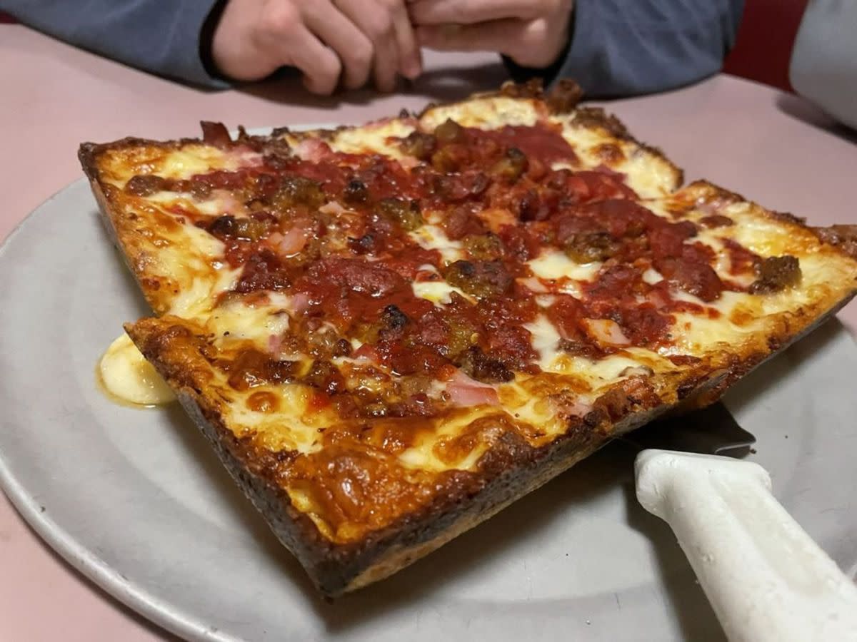 Small Meat Lovers Detroit-Style Pizza on a round pizza tray with serving spatula underneath it, Loui’s Pizza, Hazel Park, Michigan, on a pink table with a person’s hands and arms at the top