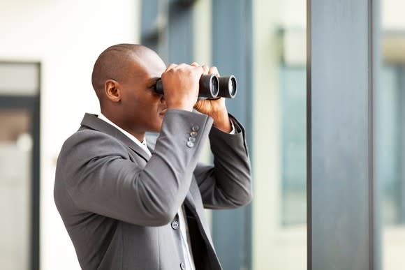 Holding binoculars to his face, a man looks out a window.