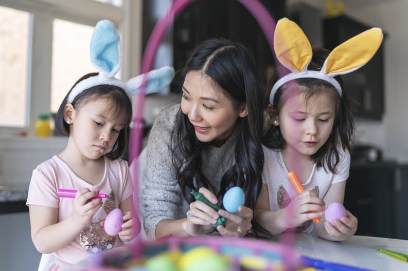 A smiling parent with two young kids wearing bunny ears while drawing on colored eggs next to an Easter basket at the kitchen table..