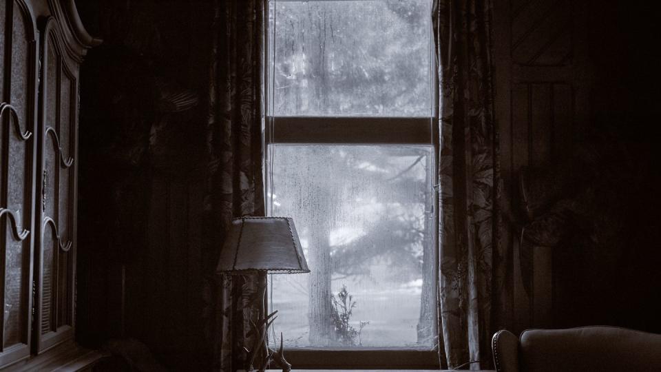 looking out of a dirty window with a curtain and a vintage lamp on the windowsill wooden living room cabinet and home interior a tree is in front of the spooky house