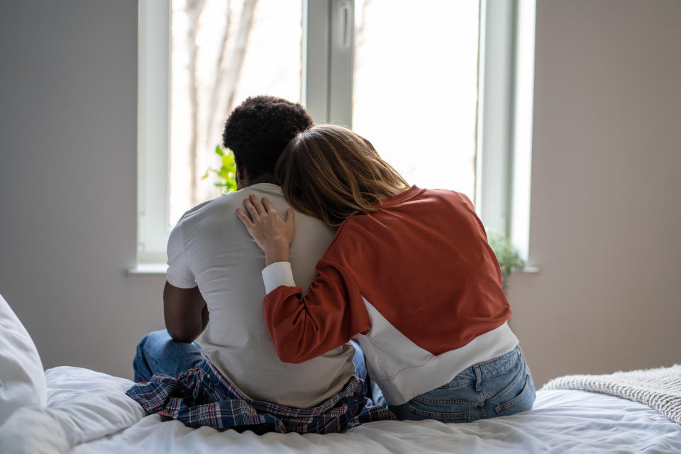 A couple sits on a bed facing a window, with the woman resting her arm on the man's back in a comforting gesture