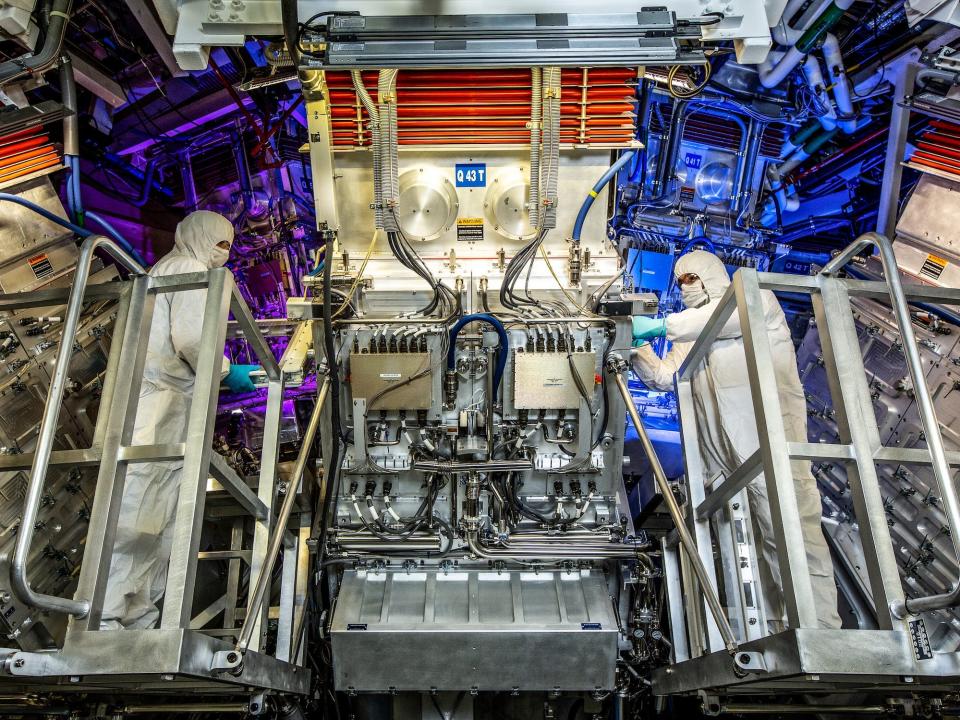 Two people in white coverall suits look at the machinery at the National Ignition Facility Target Area.