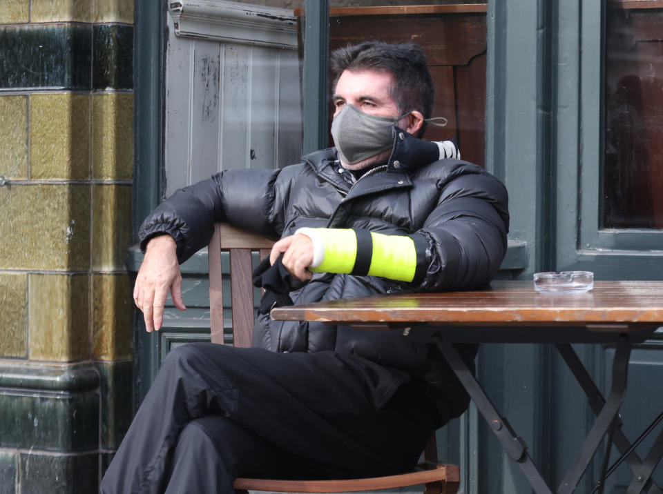 Simon Cowell sits outside The Castle pub in London's Holland Park wearing his cast