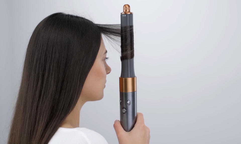 Still from a product video showing a woman holding the Dyson Airwrap styling tool vertically as it wraps her long black hair around its barrel. Gray background.