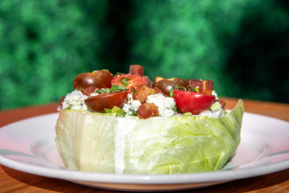 A wedge salad on a white plate, featuring iceberg lettuce, grape tomatoes, blue cheese crumbles, bacon bits, and chives, on a blurred green background