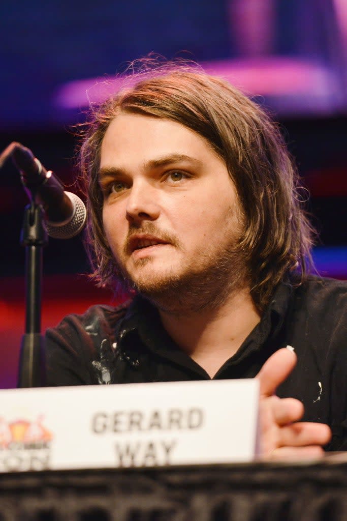 Gerard Way speaking at a panel, seated behind a sign with his name. He is wearing a jacket with shoulder details