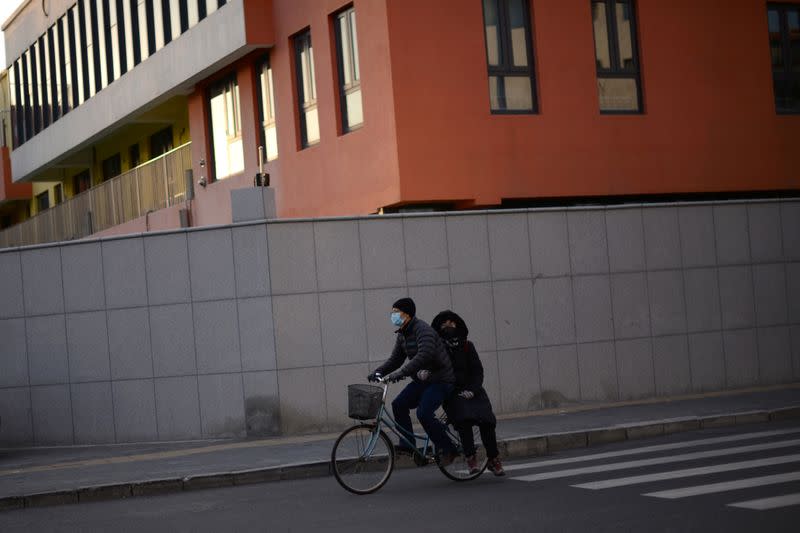 A man wearing a face mask rides a bicycle with a passenger on the back, in Beijing