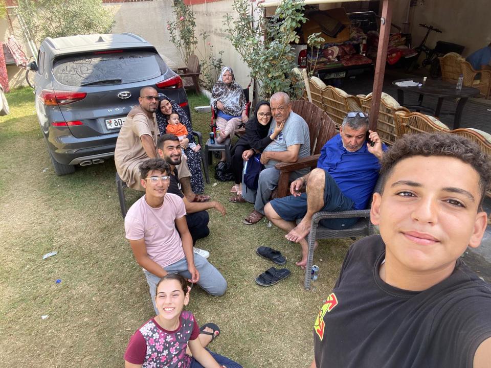 A selfie shows a large family relaxing near a parked car.