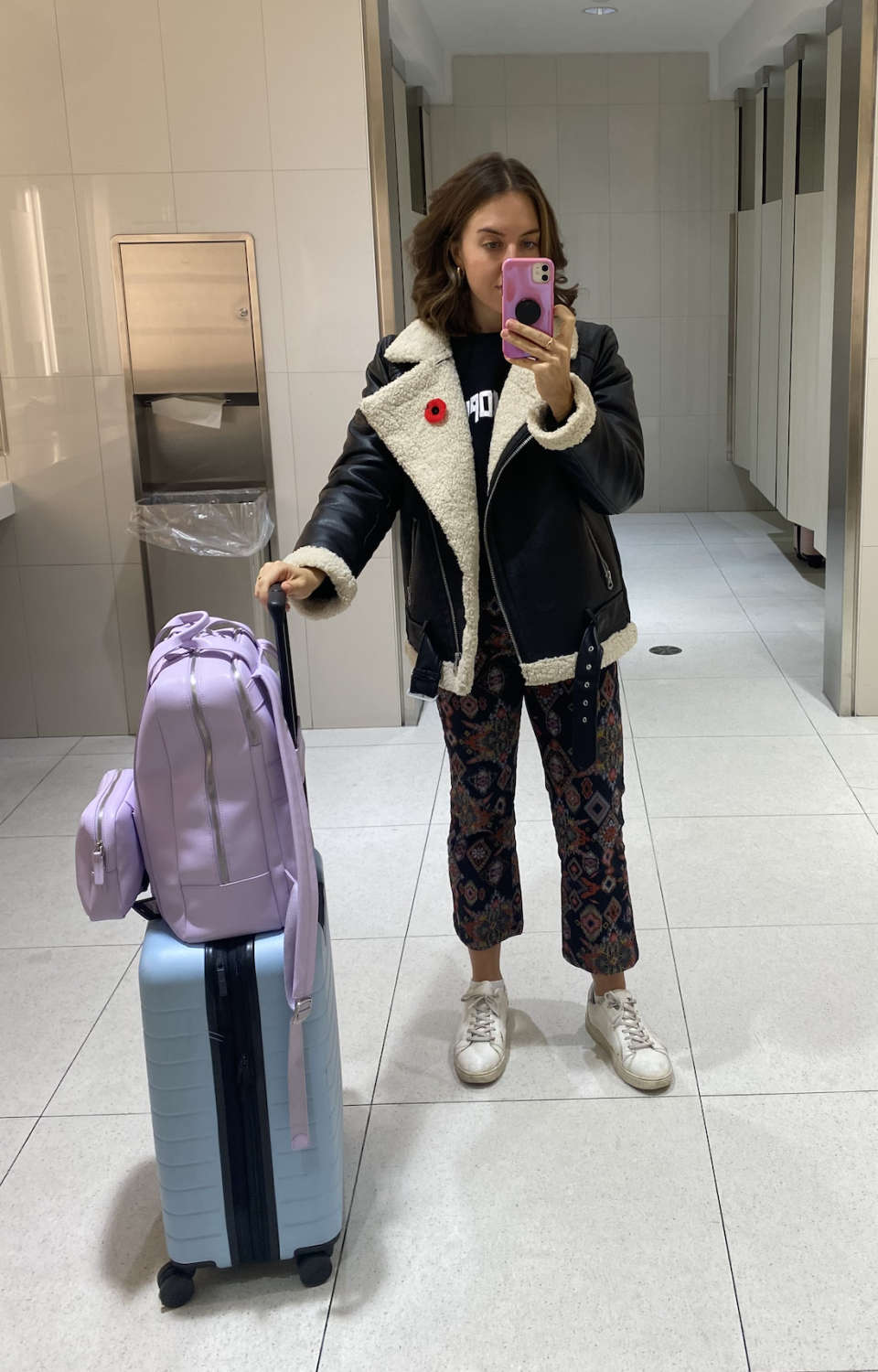 brunette woman taking mirror selfie with blue suitcase, purple backpack, white sneakers, printed pants, black aviator jacket