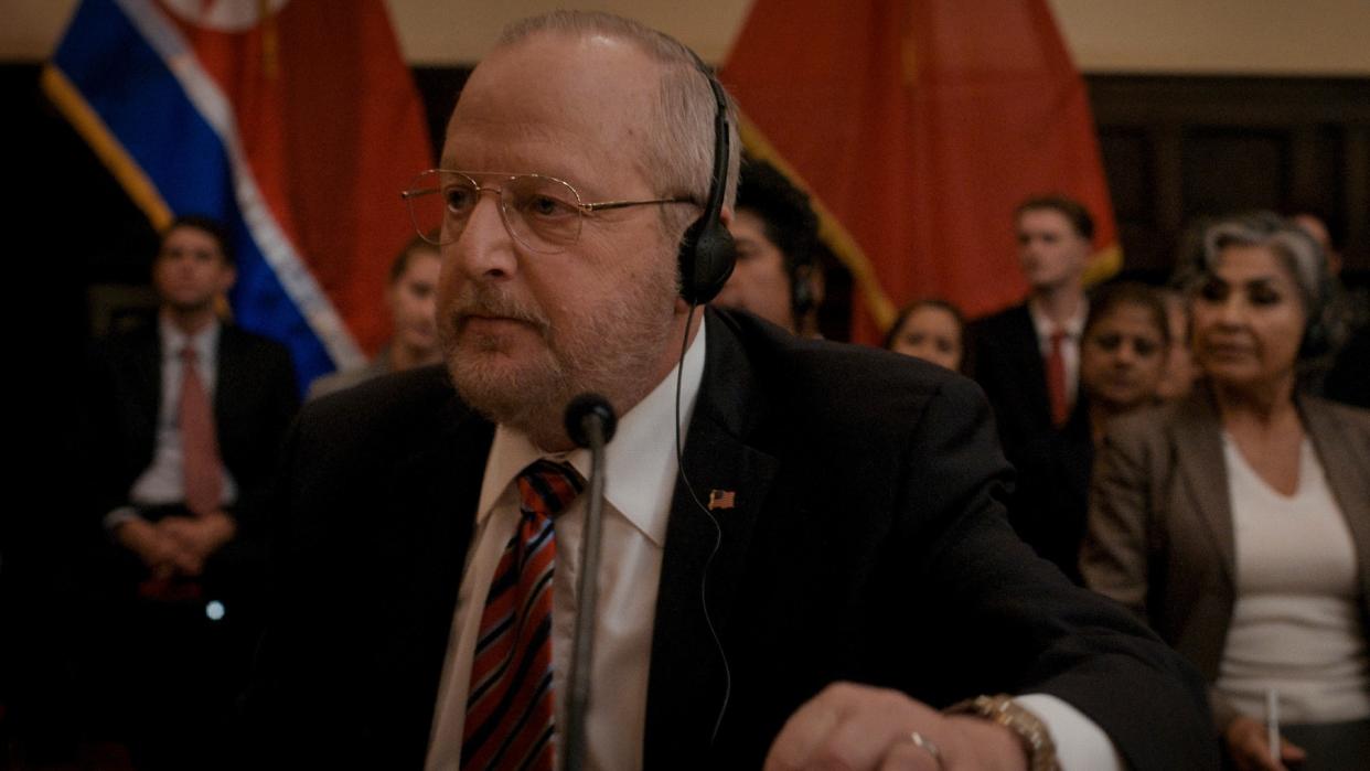  Close up of an older man speaking into a microphone at a big conference. He has short gray hair, beard, and moustache. He is wearing gold glasses and some black headphones. The man is dressed in a dark suit, white shirt, and red and blue striped tie. On his jacket lapel there is a small American flag pin. In the background you can see lots of well-dressed people sitting down, listening, as well as some official looking flags. 
