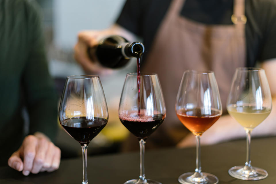 Person pouring wine into a glass with other glasses containing different types of wine lined up on a counter