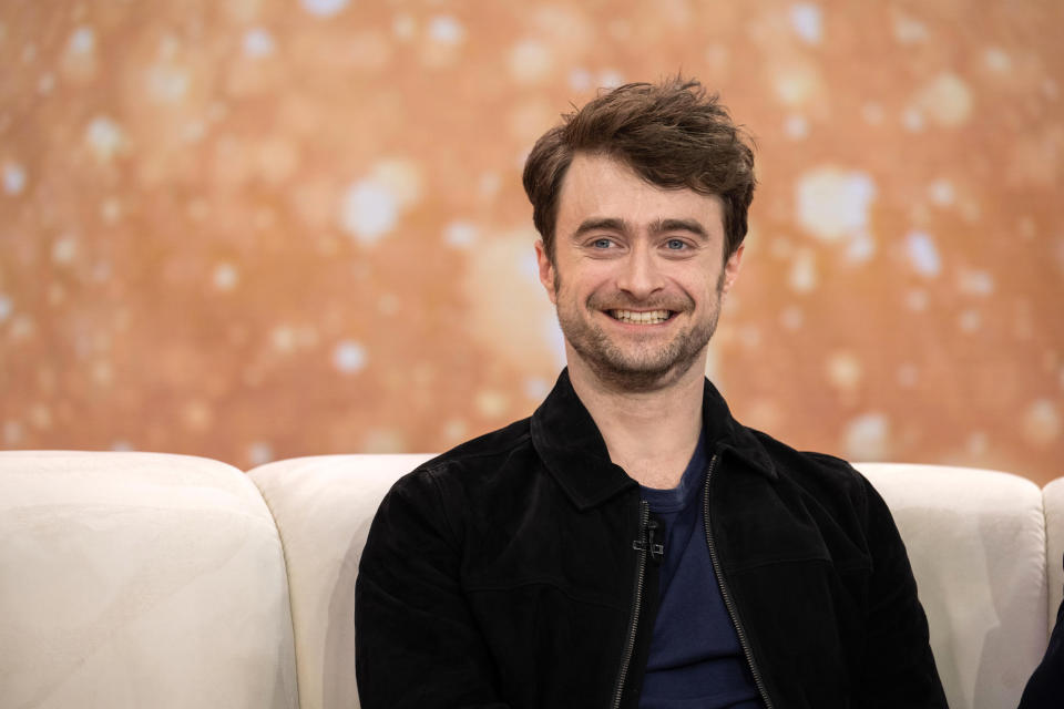 Daniel Radcliffe smiling while seated on a white couch during an interview