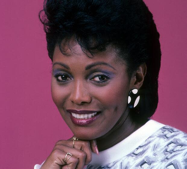 A Black woman with short hair, blue eyeshadow and large earrings in a patterned shirt posing with her hand under her chin