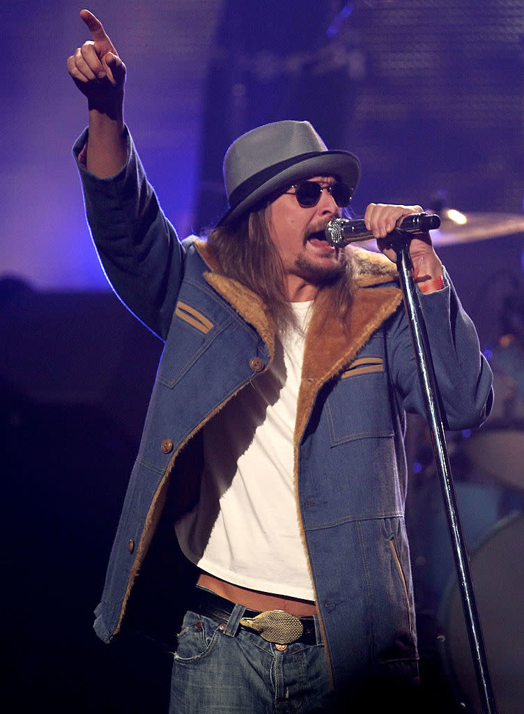 Kid Rock performs on stage, wearing a hat, sunglasses, a fur-lined jacket, and jeans, singing into a microphone while pointing with his other hand