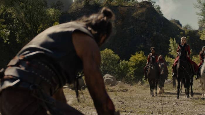 A man in a leather outfit and top-knot faces down four mounted Red Ajah Aes Sedai (led by Liandrin, played by Kate Fleetwood) who have cornered him