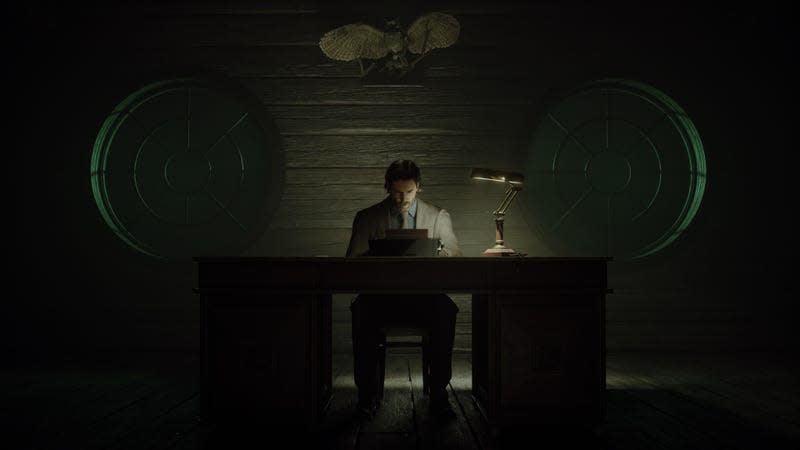 Alan Wake sits at a desk in front of a typewriter, in a dark room with two round windows behind him.