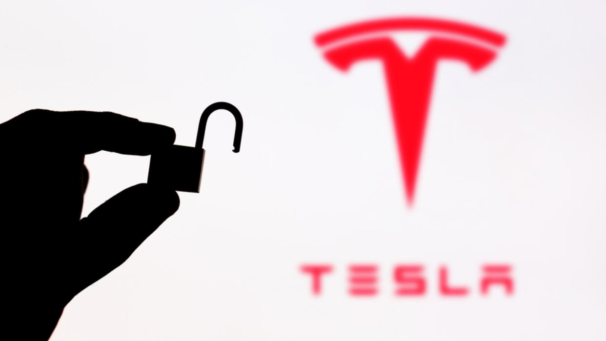  A hand holding an unlocked padlock in front of a blurry tesla logo. 