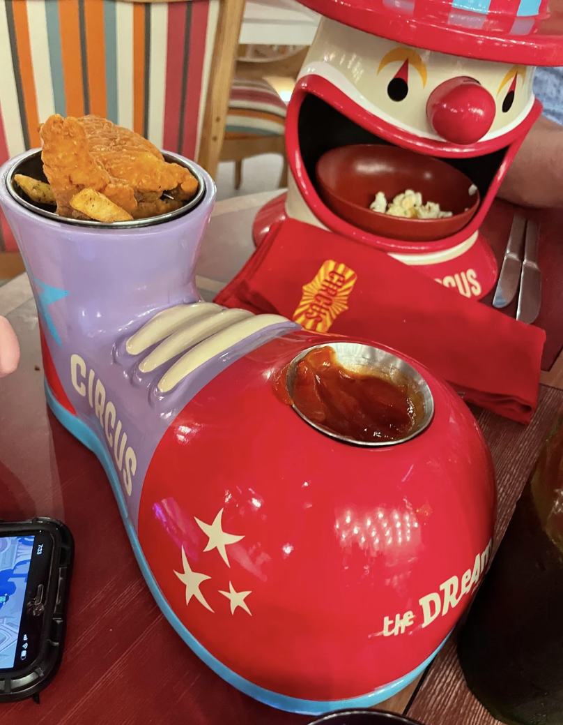 A novelty circus-themed serving dish with a clown face and shoe, holding chicken tenders and sauce