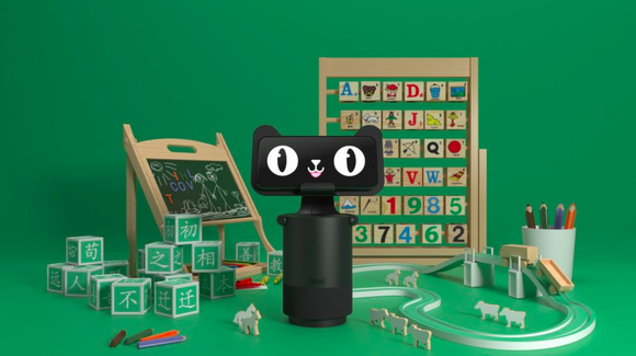 Alibaba's smart speaker, the Tmall Genie, is pictured with its new visual component and surrounded by educational toys against a green backdrop