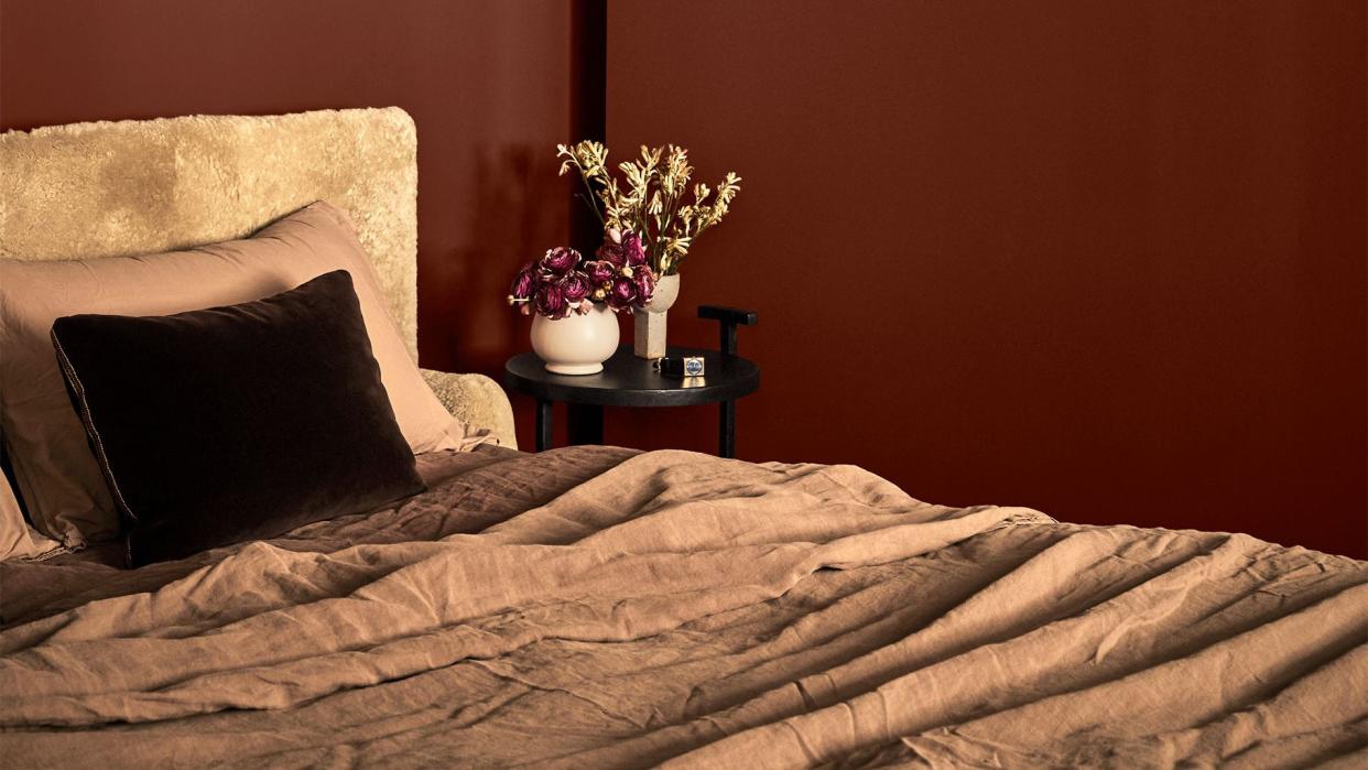 in a brown painted bedroom, a bed's headboard is covered in a light brown shearling, bed covering is in a medium brown linen, a small round night table has a plant and a vase with flowers, a small artwork on wall