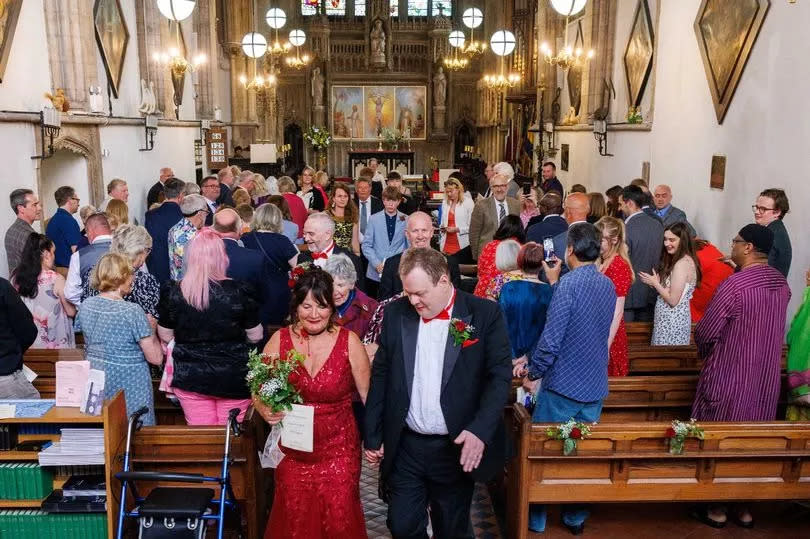 Couple in wedding outfits walk down aisle as wedding guests look on from pews in chapel