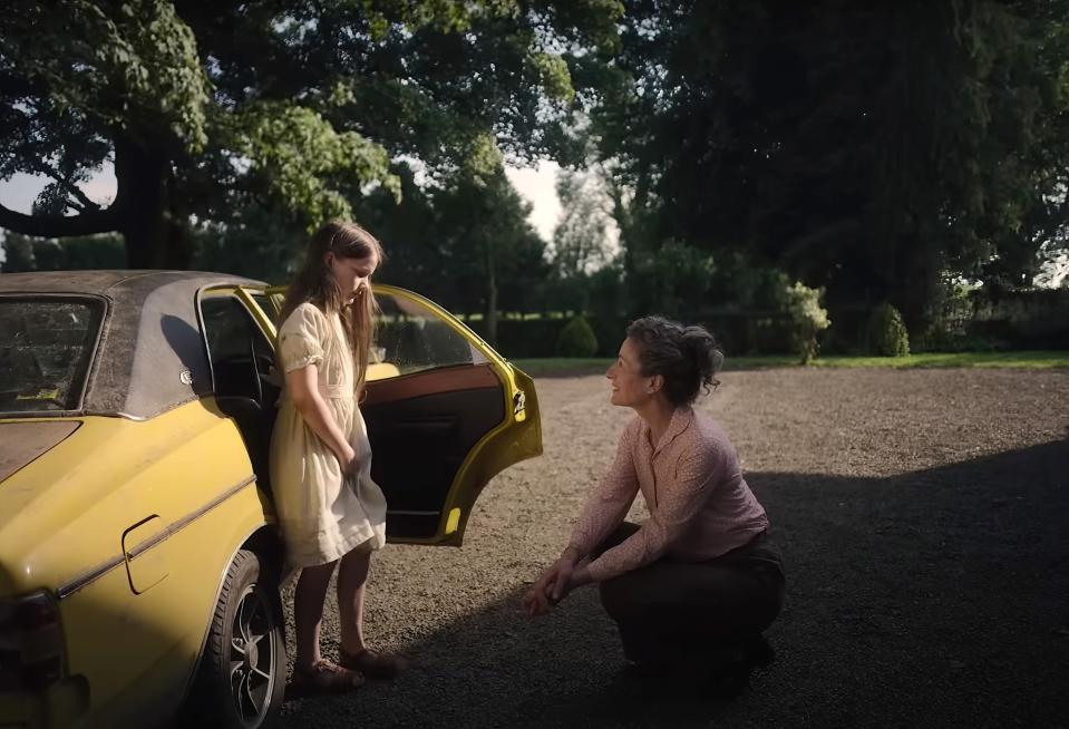 A woman kneels to talk to a young girl standing by an open car door