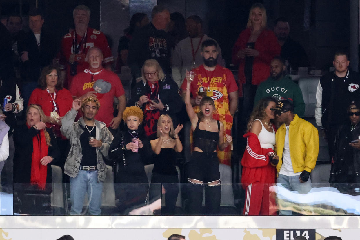 Taylor Swift, surrounded by several dozen others in a stadium box, holds up a glass.