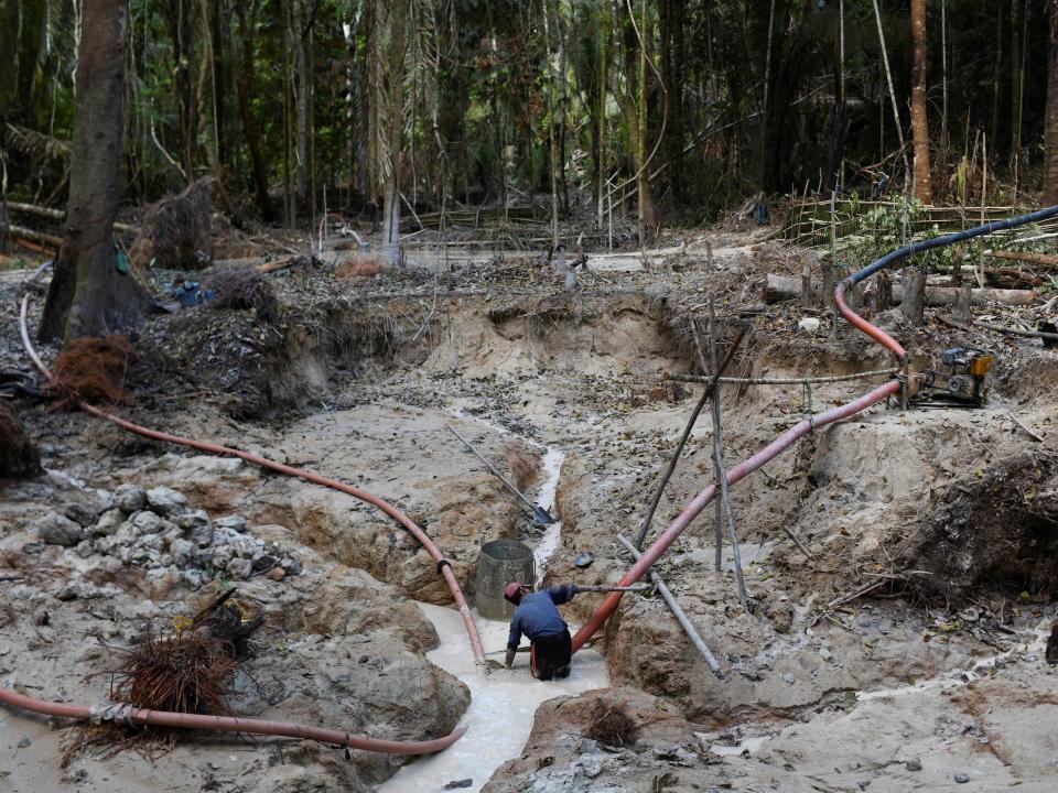 man stands in muddy pit with pipes trailing away in every direction in cleared and dredged area of amazon rainforest