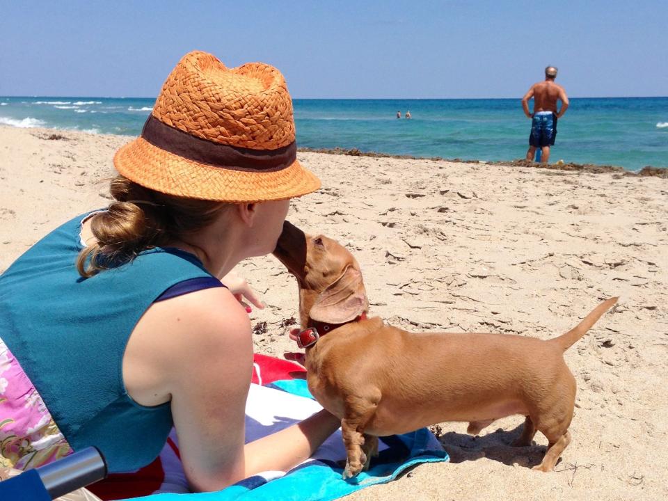 A woman wearing a hat appears to kiss her dachshund dog on the beach.