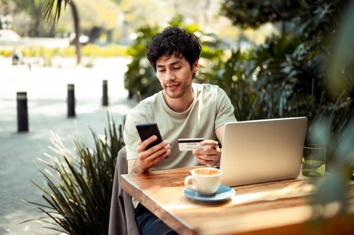 A person sitting at an outdoor cafe table holding a credit card and phone with open in front of them.