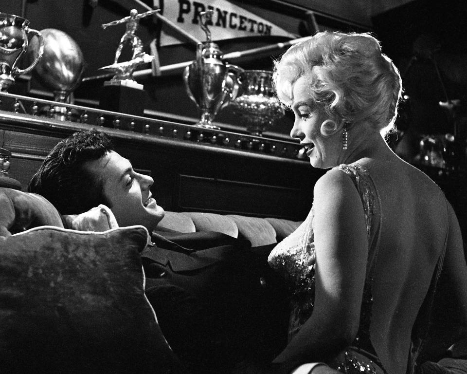 Tony Curtis and Marilyn Monroe in a scene from the movie "Some Like It Hot," with Curtis lying on a couch and Monroe wearing an elegant, sleeveless dress