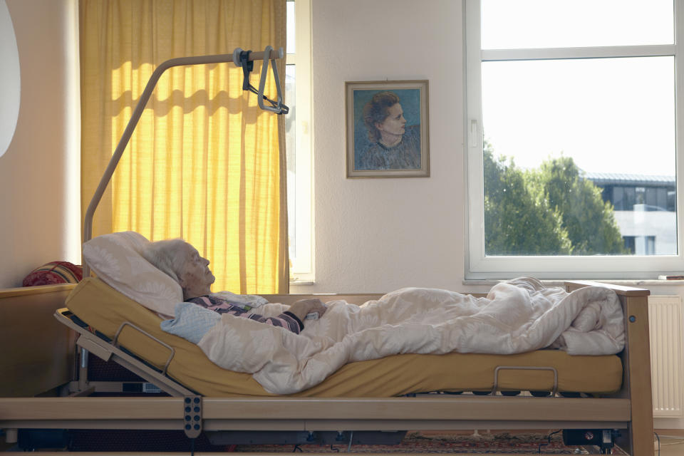 Elder person resting in a hospital bed by a window with a portrait on the wall