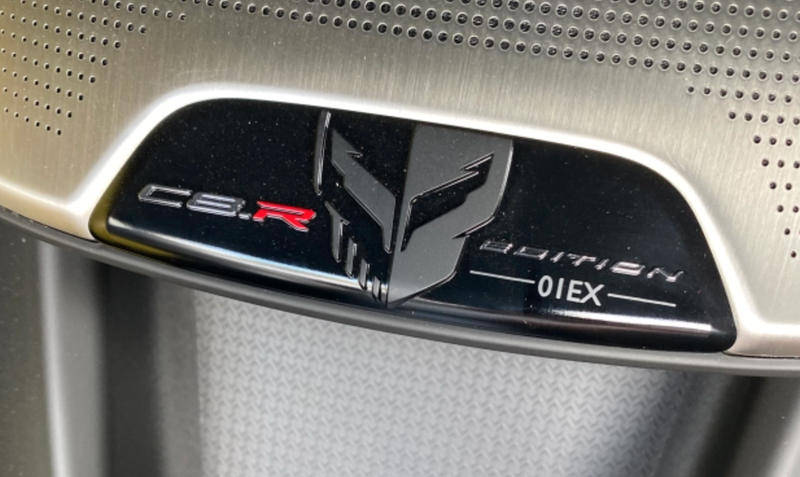 Close-up photo of a commemorative plaque on the dashboard of a 2021 Chevy Corvette. The plaque reads "C8.R Edition - 01EX," with the Corvette Racing skull logo in the center.