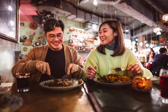 Two people enjoy a meal together in a cozy restaurant, smiling and chatting. The table is set with dishes and drinks.