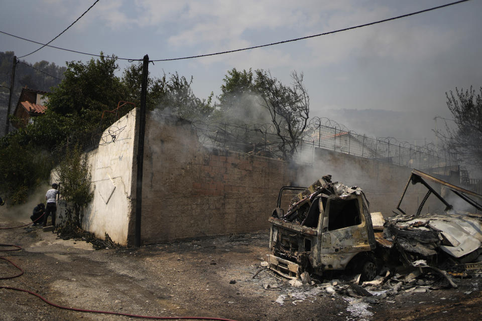 Two men try to control a wildfire in a house as smokes rises from a burn truck in Acharnes, a suburb of northern Athens, Greece, We dnesday, Aug. 23, 2023. Water-dropping planes from several European countries joined hundreds of firefighters Wednesday battling wildfires raging for days across Greece that have left 20 people dead, while major blazes were also burning in Spain's Tenerife and in northwestern Turkey near the Greek border. (AP Photo/Thanassis Stavrakis)