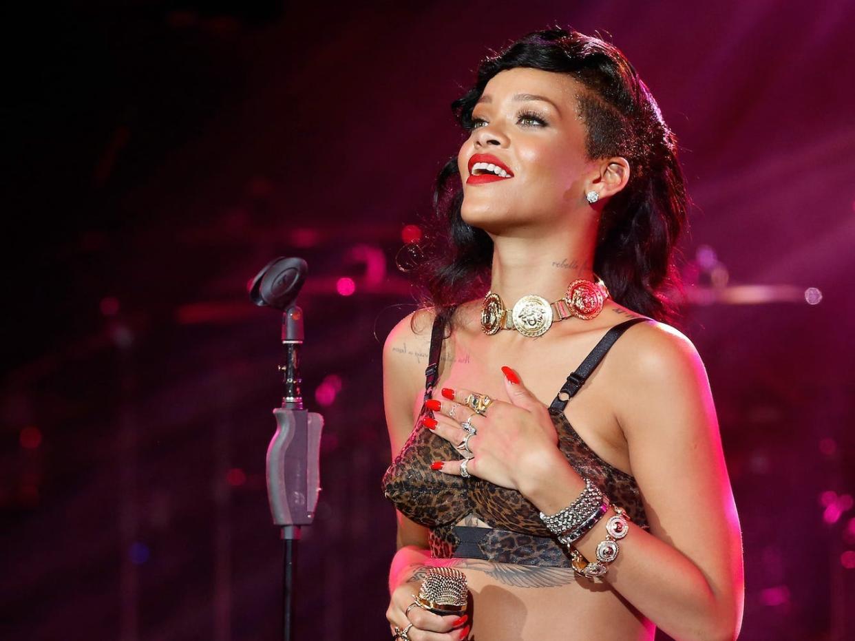 A woman (singer Rihanna) stands with one hand on her chest and a microphone in the other hand, smiling out into a crowd at her concert.