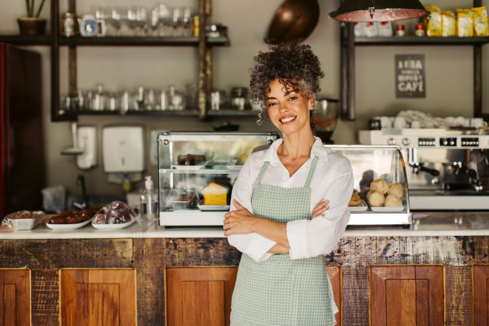 Smiling business owner standing with her arms crossed. Successful mature businesswoman feeling confident while wearing an apron in front of her coffee shop during the day.