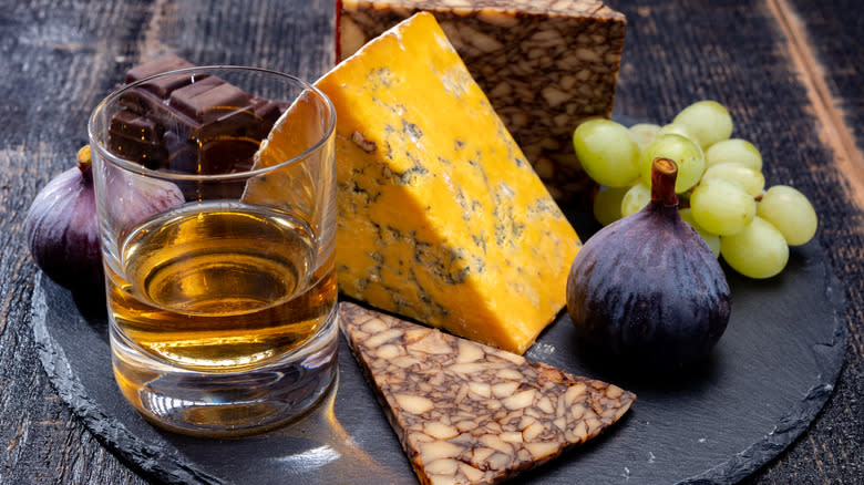 A glass of neat whiskey next to cheese, figs, and grapes