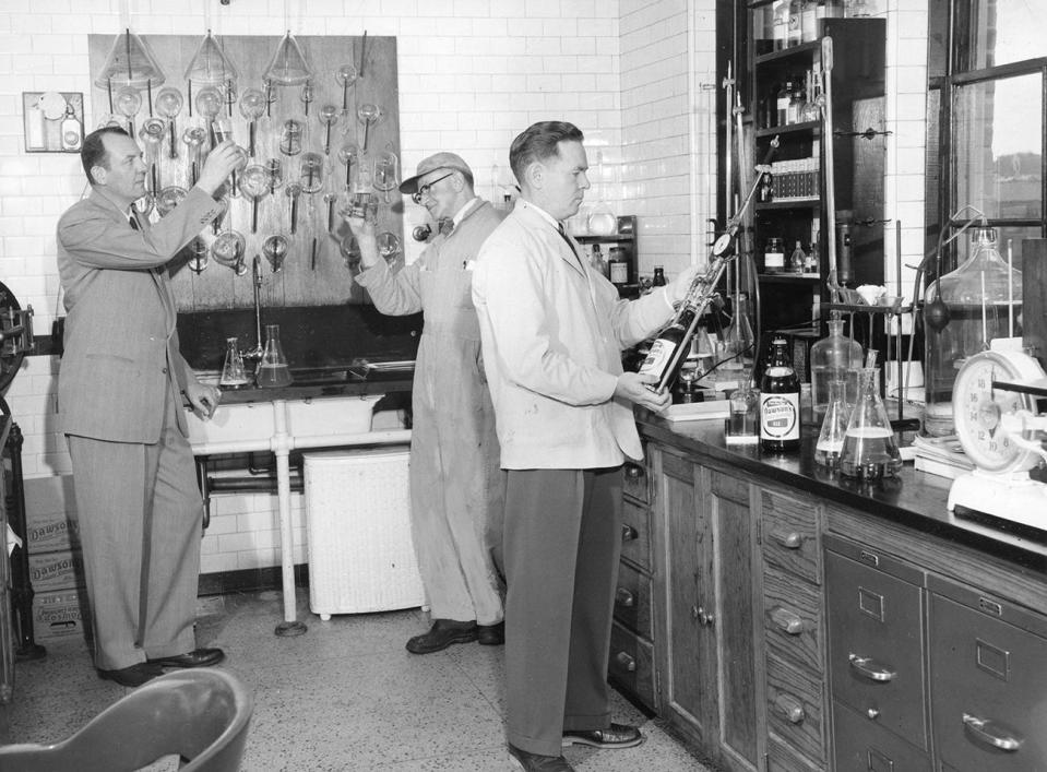 Brewmaster Theodore A. Schueler and his assistant, Herman Anderson, observes and discuss a sample of beer as Thomas McFadden, plant chemist, makes a routine test on the finished product.
(Credit: Standard-Times via Spinner Publications)