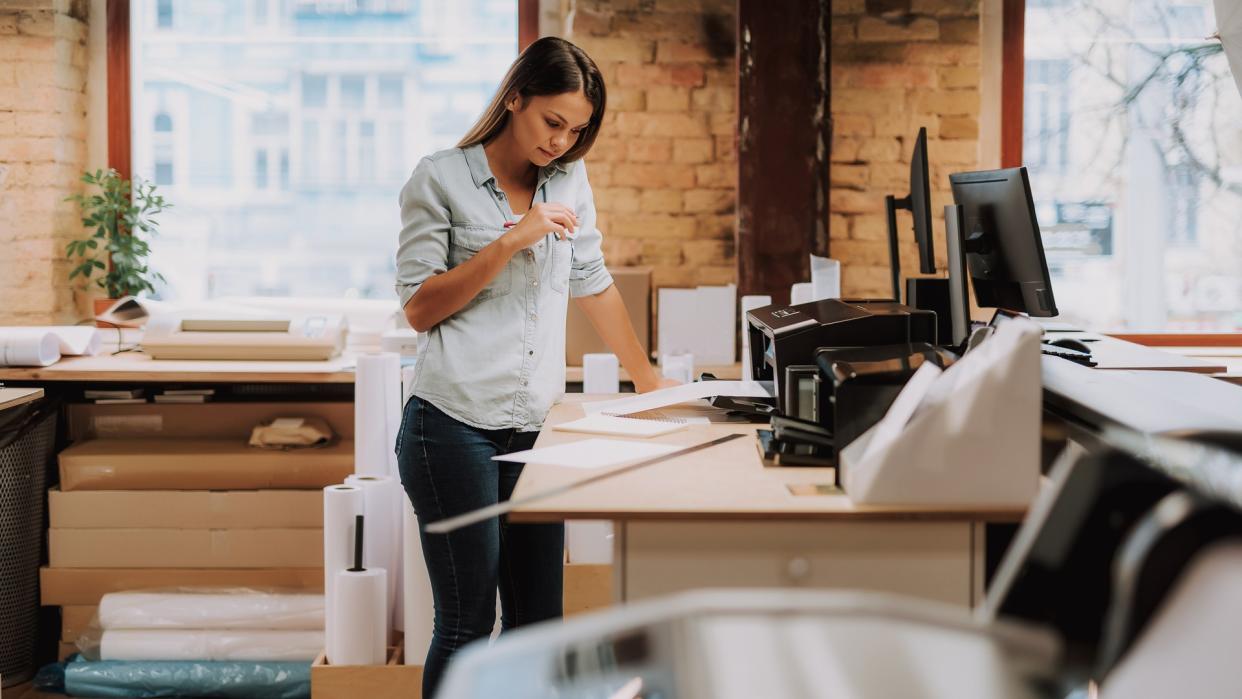 Portrait of charming woman in blue shirt looking at notes while standing near table with printer.