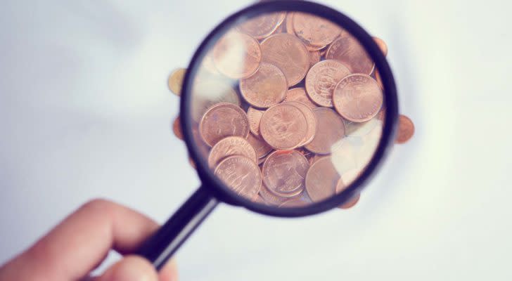 A hand holding a magnifying glass over pennies. under-$1 stocks to buy