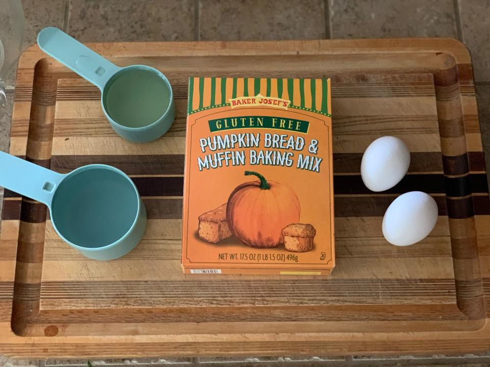 orange box of trader joe's gluten free pumpkin bread on a wooden cutting board with two measuring cups and eggs