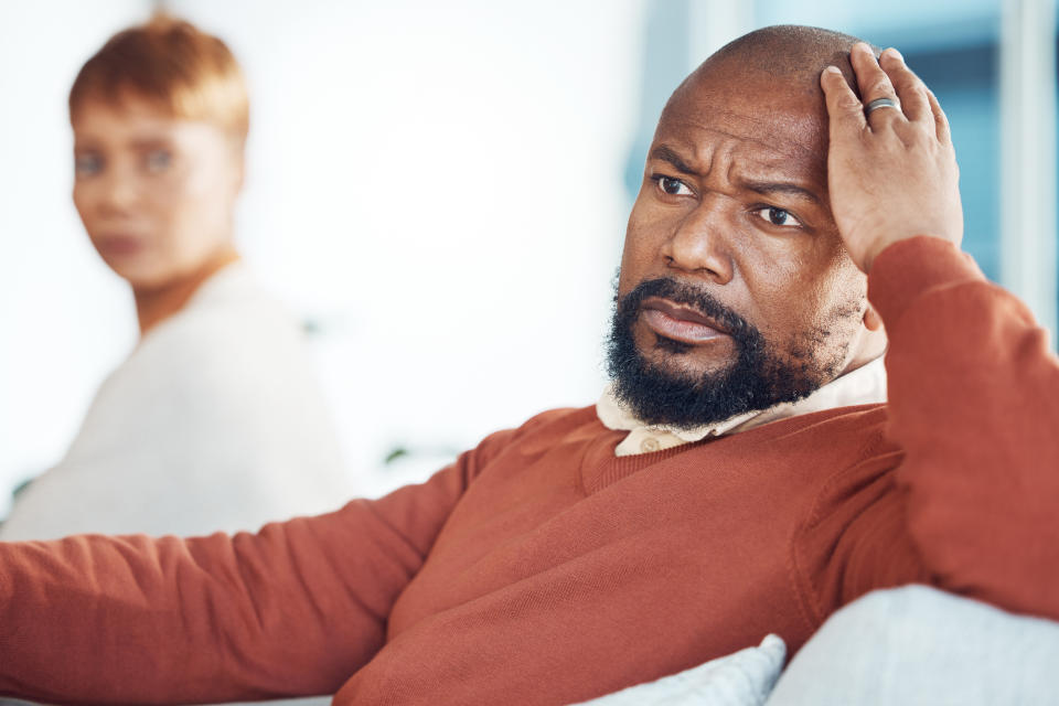 A man looks distressed, holding his head, while a woman in the background looks at him with concern. Both are casually dressed